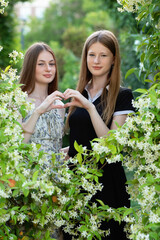 Two teenage sisters stand among blooming bushes, forming a heart with their hands. A tender portrait of sisterly love, friendship, and connection in nature’s embrace