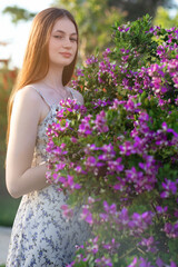 Young woman in a floral dress stands beside a blooming bush with vibrant purple flowers. A serene portrait capturing natural beauty, youth, and spring freshness.