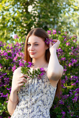 Young woman in a floral dress holds purple flowers and tucks one in her hair, surrounded by blooming bushes. A fresh, joyful portrait full of spring charm