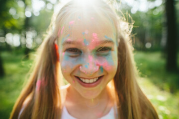 Caucasian girl smiles with colorful powder on face under forest light. Childhood joy, authenticity, emotional freedom and self expression in natural summer setting.
