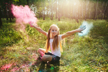 Caucasian girl sits in grass and throws pink and blue powder in sunlit forest. Concept of playful...