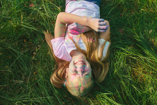 Overhead view of happy white girl with long hair lying on grass, laughing with eyes closed, face and arms stained with colorful powder. Concept of playful childhood, authentic joy, creativity, and emo