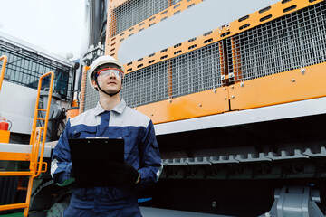 Caucasian male engineer overseeing machinery in mining industry with clipboard and protective gear