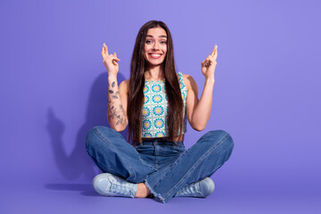 Smiling young woman with crossed fingers sitting on violet background showcasing cheerful and...