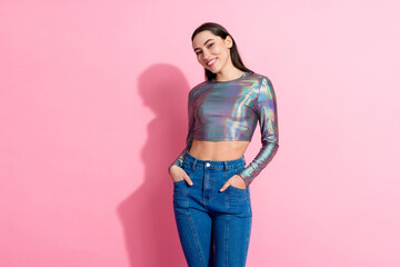 Smiling young woman in a vibrant glossy top standing confidently against a bright pink background with hands in jeans pockets