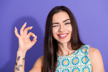 Smiling young woman making an OK hand gesture on a purple background dressed in colorful printed...
