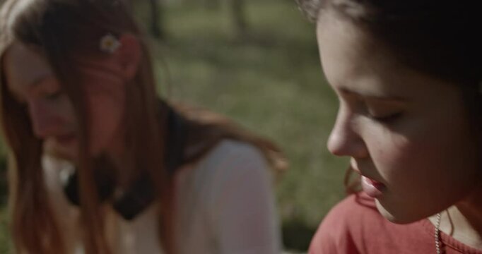 Two teenage girls enjoy a beautiful day in the park. They sit on a blanket in the grass, talking and drinking water. One girl adjusts a scrunchie in her friend's hair.