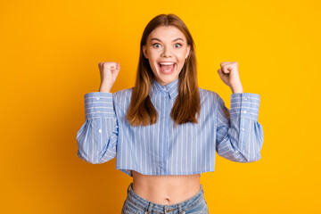 Excited young woman in blue shirt with fists raised celebrating success against yellow background