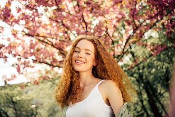 Joyful young woman with curly red hair smiles outdoors amidst blooming trees during a sunny spring...