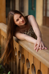 Smiling young woman with long, shiny, well-groomed hair leans over a wooden railing. A beautiful example of healthy hair care, softness, and natural beauty