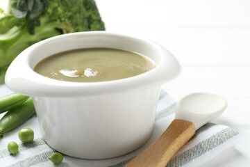 Baby food. Tasty puree in bowl, spoon, peas and broccoli on white table, closeup