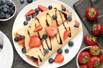Tasty crepes with strawberries, blueberries and chocolate sauce on gray textured table, flat lay