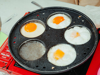 Close-up of sunny side up eggs being cooked on a multi-hole frying pan over a portable gas stove,...