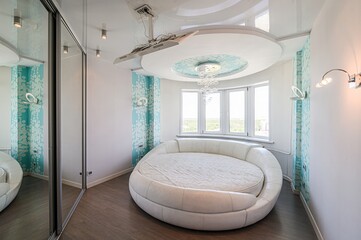 bright bedroom features a round white bed under a chandelier, by a window. A wardrobe with mirrored...