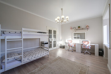 Bright room features white bunk beds, a shared desk with pink chairs, and a beige rug. A chandelier hangs from the ceiling, adding a touch of elegance