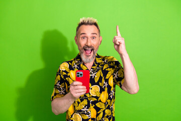 Charismatic mature man with a funky blond haircut wearing a summer print shirt, holding a red smartphone, on green background