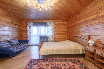 Cozy wooden bedroom with a bed, couch, patterned rug, and window. Warm lighting and a chandelier...