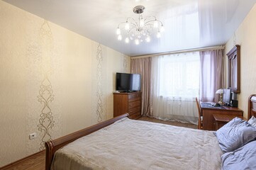 cozy bedroom with a double bed, wooden furniture, patterned wallpaper, and a chandelier. A desk is...