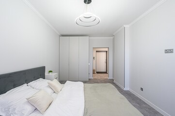 bright, modern bedroom features a bed with white linens and a gray headboard. A wardrobe stands near an open doorway, with neutral tones throughout the space