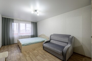 cozy room features a bed with a light blue patterned cover, a grey sofa, and a window with grey curtains. The floor is wooden-like