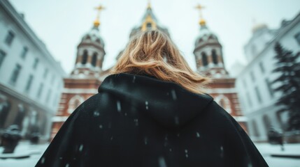 A woman stands in snow, facing a beautiful church with domes, lost in thought and reflection amidst the serene winter landscape.