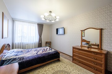 cozy bedroom with a bed, wooden dresser with a mirror, a TV, and patterned wallpaper. Soft light...