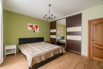 Cozy bedroom features an olive green wall, a bed with a patterned quilt, a wardrobe with mirrored...