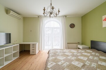 cozy bedroom with light green walls features a bed, desk, TV stand, and a large arched window with...