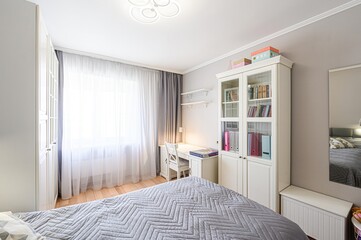 bright, tidy bedroom with light walls, a large window with sheer curtains, white furniture, and a bed with a grey patterned duvet