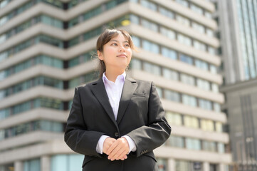 スーツ姿で穏やかな表情を見せる新入社員の女性がビル街に立つ