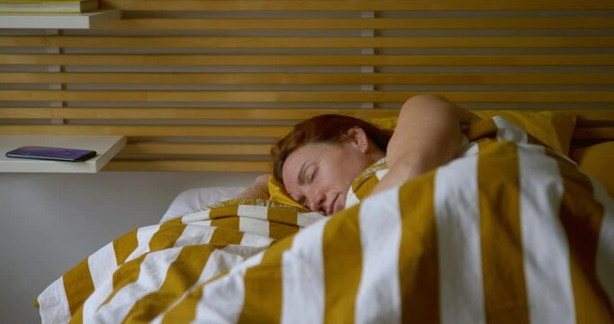 Adult woman sleeping in bed in her room, the alarm clock on the mobile phone on the bedside table rings, she wakes up, picks it up, looks at it and stretches. In the morning