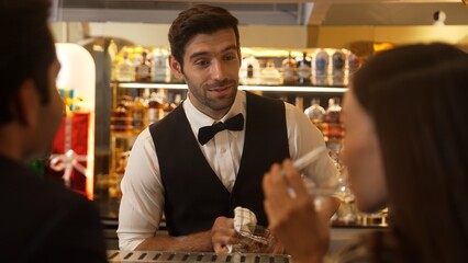 Focusing smiling smart bartender serving gin fizz or tonic soda at luxurious bar, engaging lively conversation with VIP customer couple at back view in atmosphere at lighting warm nightclub. Vinosity.