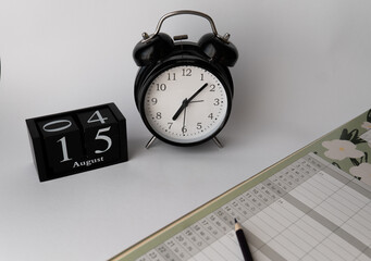 Top view of a classic black alarm clock and a wooden cube calendar with the date "15" placed on a bedside table or desk. Part of the bed with a pillow and blanket is visible in the lower right corner,