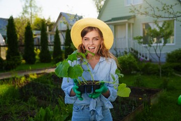 Portrait of a smiling gardener woman standing next to her green garden holding a young seedlings...