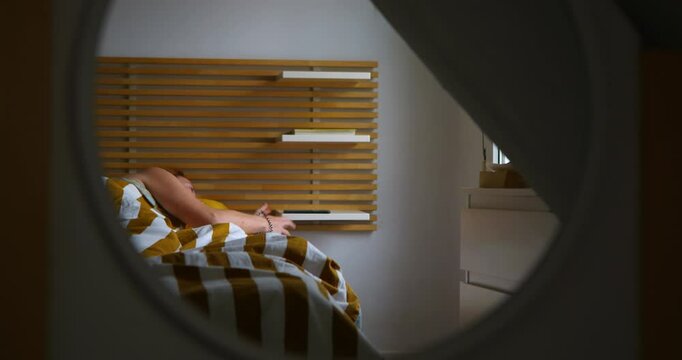 A woman reflected in a mirror is in her bedroom bed sleeping, in the morning. The alarm clock on her mobile phone rings, she wakes up and picks it up.