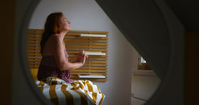 A woman reflected in a mirror is sitting on the bed in her room waking up in front of the window, stretching. In the morning.