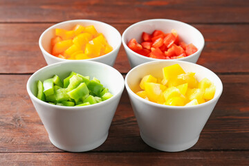 Pieces of fresh colorful bell peppers in bowls on wooden table, closeup
