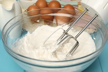 Hand mixer with whisks, whipped cream in bowl, eggs and milk on light blue table, closeup