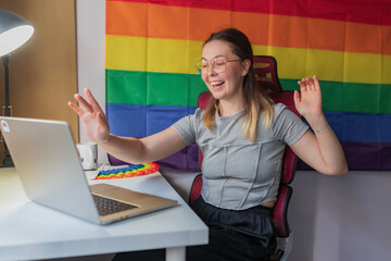 Happy woman greeting during video meeting, rainbow flag backdrop visible/	Smiling young woman...