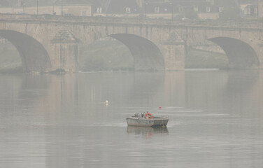 Gentle morning mist blankets the serene Loire River as a small boat glides through the calm waters,...