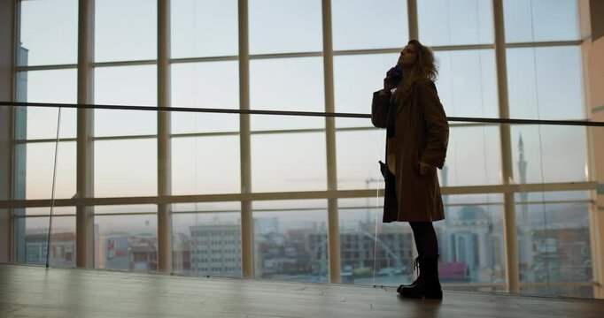 Young Woman Talking on Phone While Walking in Modern Building with City View of Pristina, Kosovo