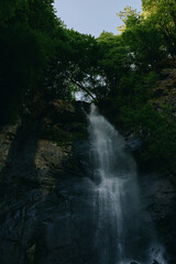 Waterfall cascading down rocky cliff surrounded by lush green forest under clear sky, creating a...