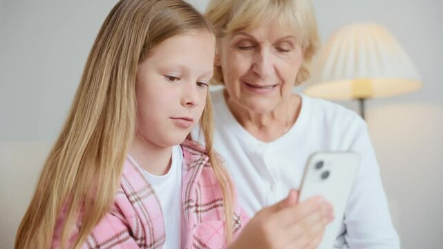 A 10-year-old girl laughs during a video call while her 60-year-old grandmother looks at the screen with a serious expression. Generational contrast in shared digital moment. High quality 4k footage