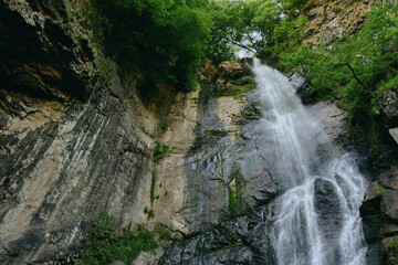 Waterfall cascading down rocky cliffs surrounded by lush green vegetation under bright daylight in...