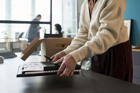 Senior Caucasian woman packing personal belongings and office supplies into cardboard box on desk, preparing for retirement in modern workspace, colleagues visible in background