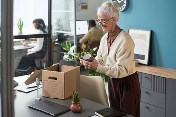 Senior Caucasian woman packing personal belongings into cardboard box in modern office, preparing for retirement while standing at desk, coworkers working in background