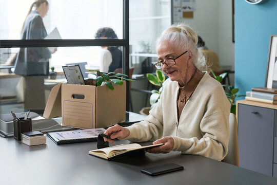 Senior Caucasian woman sitting at desk reading notebook, smiling while preparing for retirement in modern office setting, cardboard box with personal items on table, colleagues in background