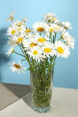 Many beautiful chamomile flowers in vase on wooden table against light blue background, closeup