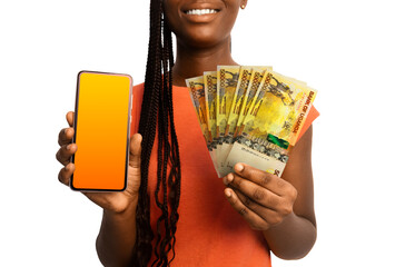 Beautiful Black woman lady smiling holding Uganda shilling notes and showing pointing phone with blank screen to the camera over white background. Hand holding Ugandan currency notes