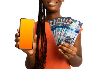 Beautiful Black woman lady smiling holding 100 Ghanaian cedi notes and showing pointing phone with blank screen to the camera over white background. Hand holding Ghanaian currency notes